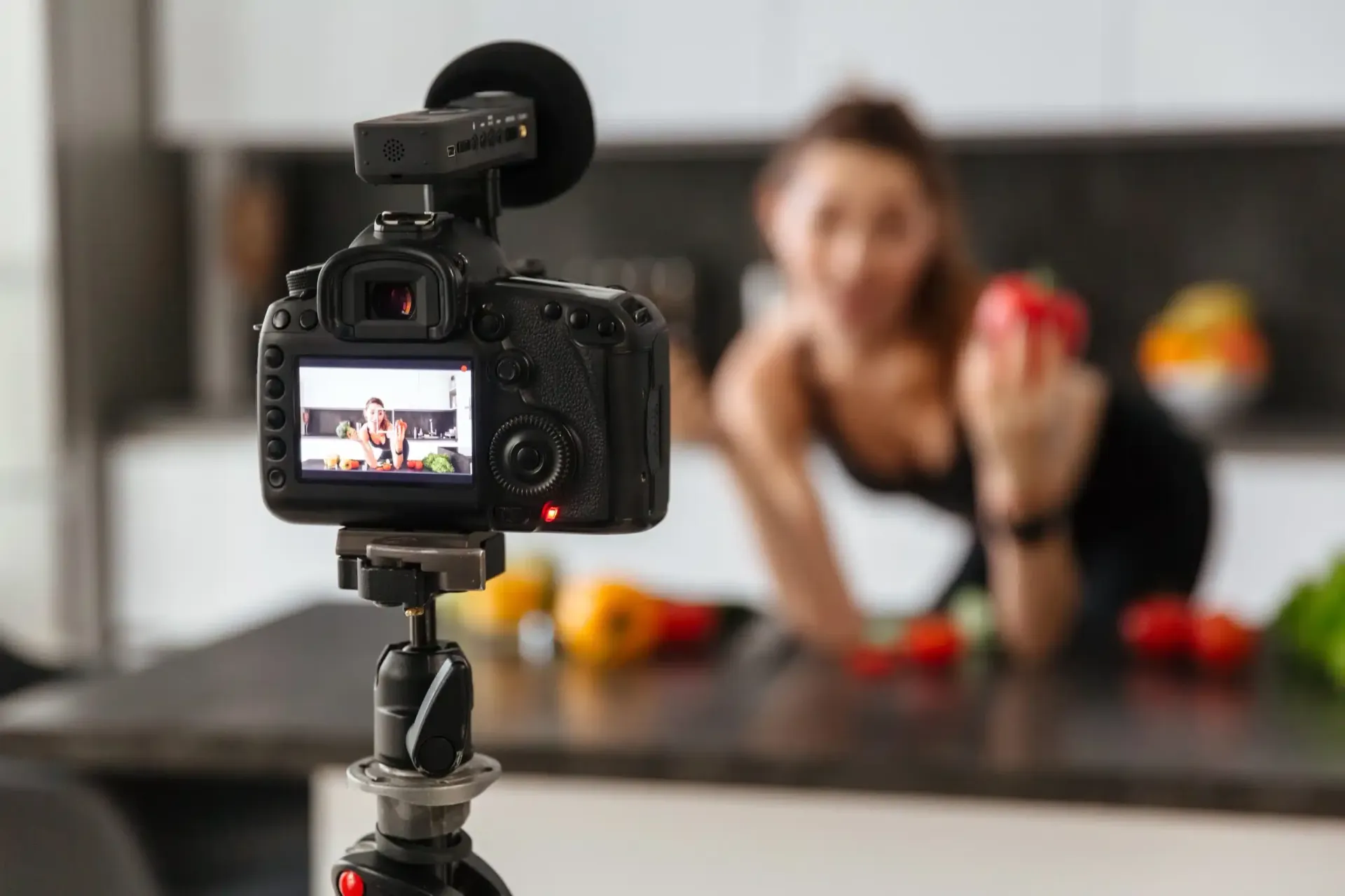 Een camera filmt een vrouw in een keuken, die een rode paprika vasthoudt. Groenten liggen op het aanrecht.