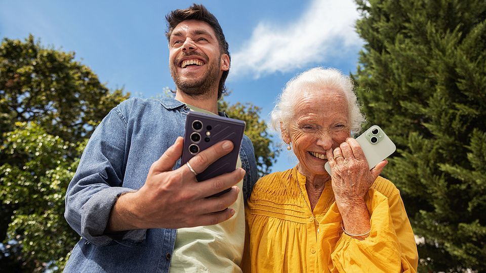 Een man en een oudere vrouw glimlachen terwijl ze buiten op hun telefoons kijken. De man houdt een paarse telefoon vast, de vrouw een witte. Het is een heldere, zonnige dag.