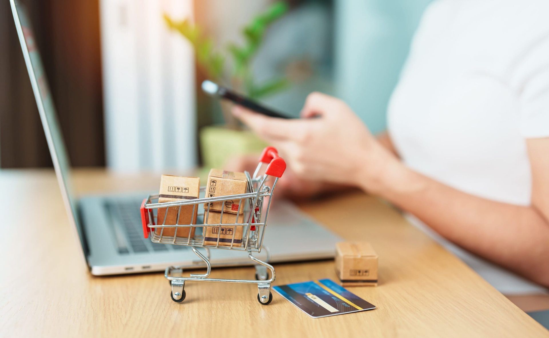 Een persoon die online winkelt met een laptop, telefoon, een miniwinkelwagentje met dozen en een creditcard op een bureau.