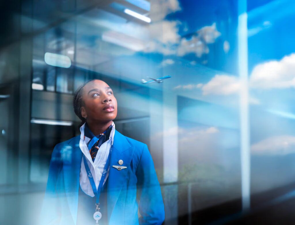 Vrouw in pyjama zit op een bed en kijkt achterom. Het bed is grijs met wit beddengoed en staat vlakbij een raam.