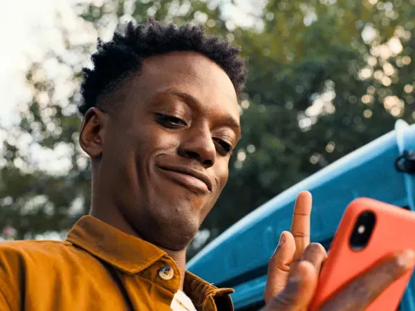 Een man met kort, donker haar grijnst naar een rode telefoon. Hij wijst met één vinger omhoog, vlakbij een turquoise voertuig.