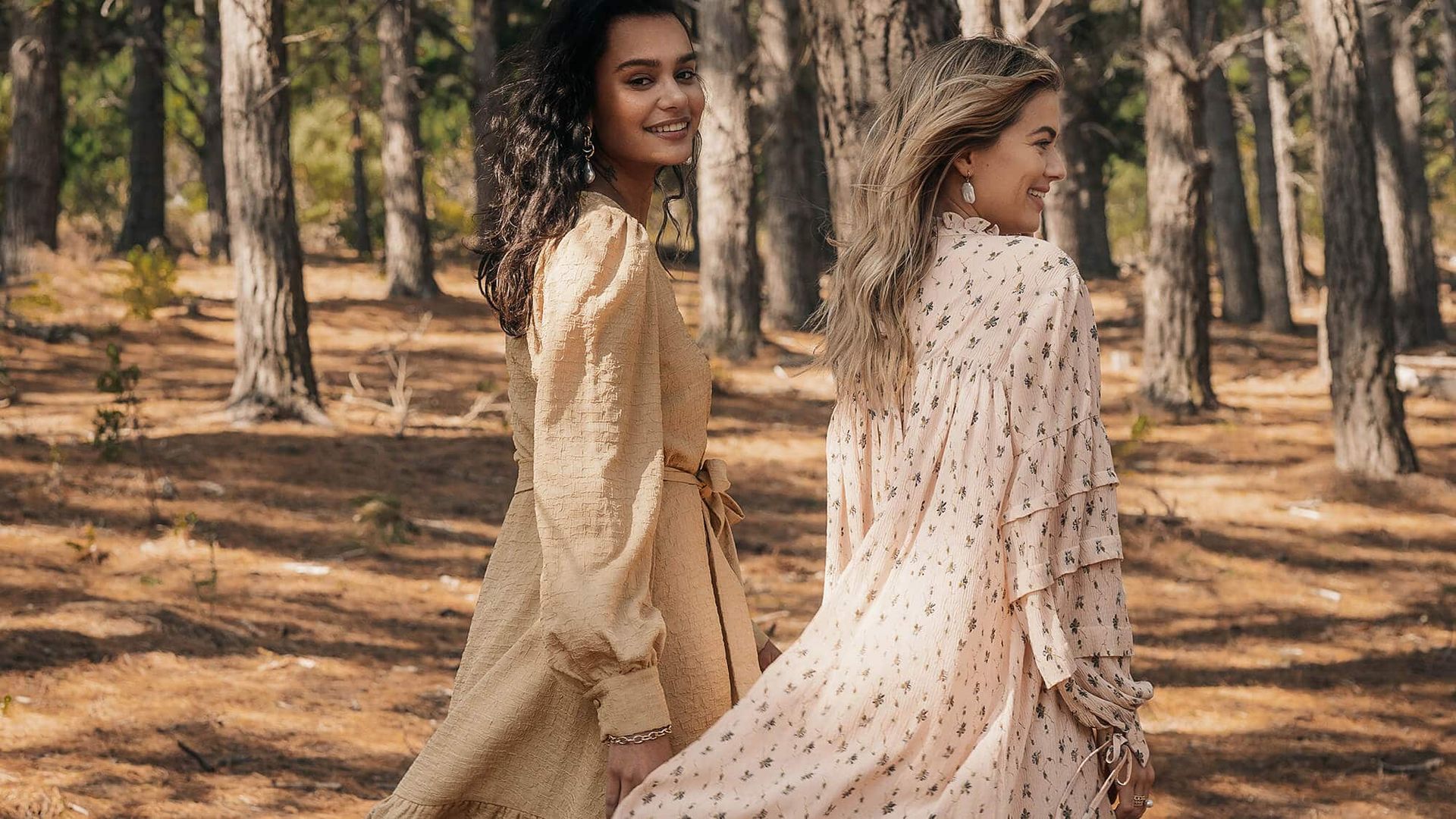 Twee vrouwen glimlachen terwijl ze in een bos wandelen. De ene draagt ​​een lichtbruine jurk, de andere een roze jurk met bloemenprint.