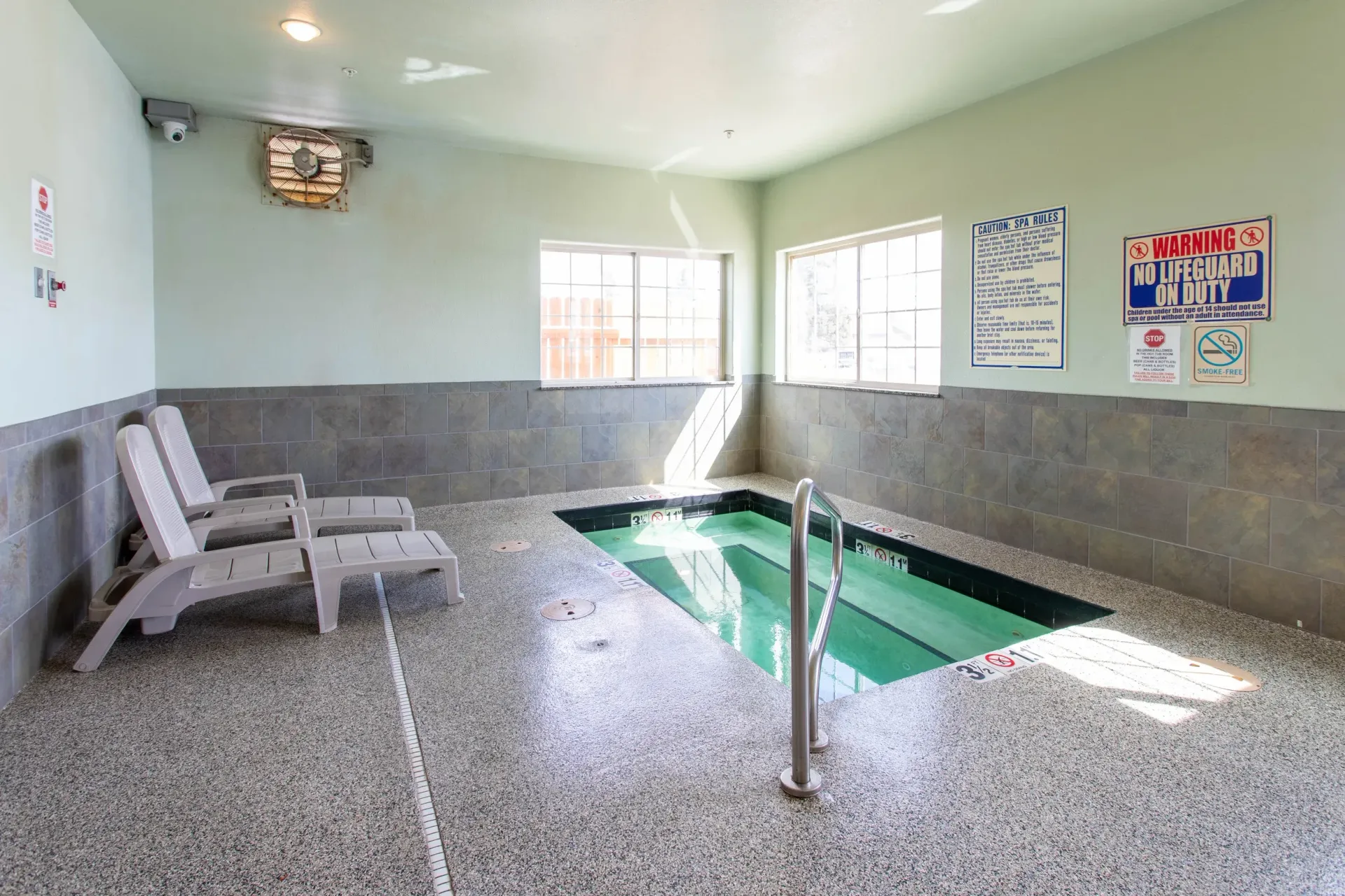 Indoor spa with lounge chairs, handrail, windows, and safety signage. Light green walls, speckled flooring.