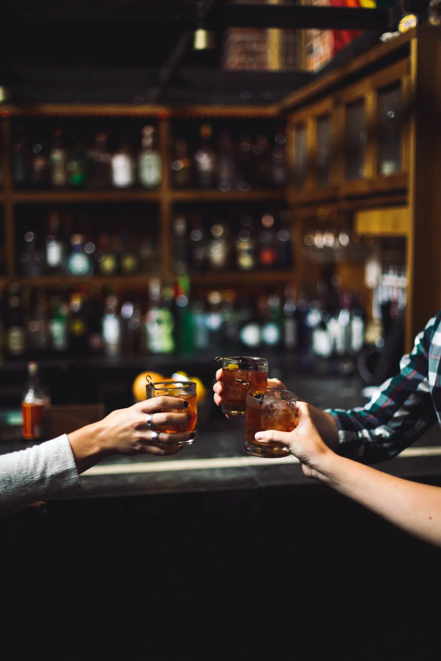 Hands clinking glasses of cocktails at a bar, dark wood backdrop, blurred bottles.