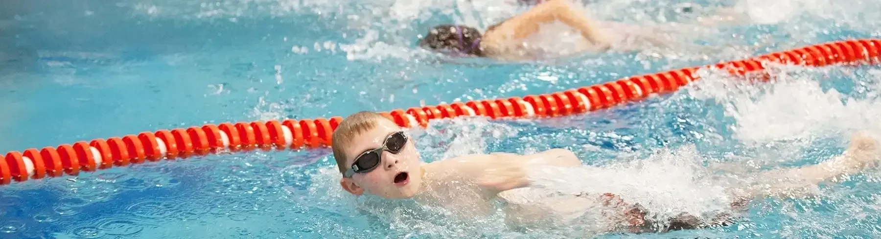 A young swimmer with goggles swims in a pool, splashing. An orange lane marker floats in the water.