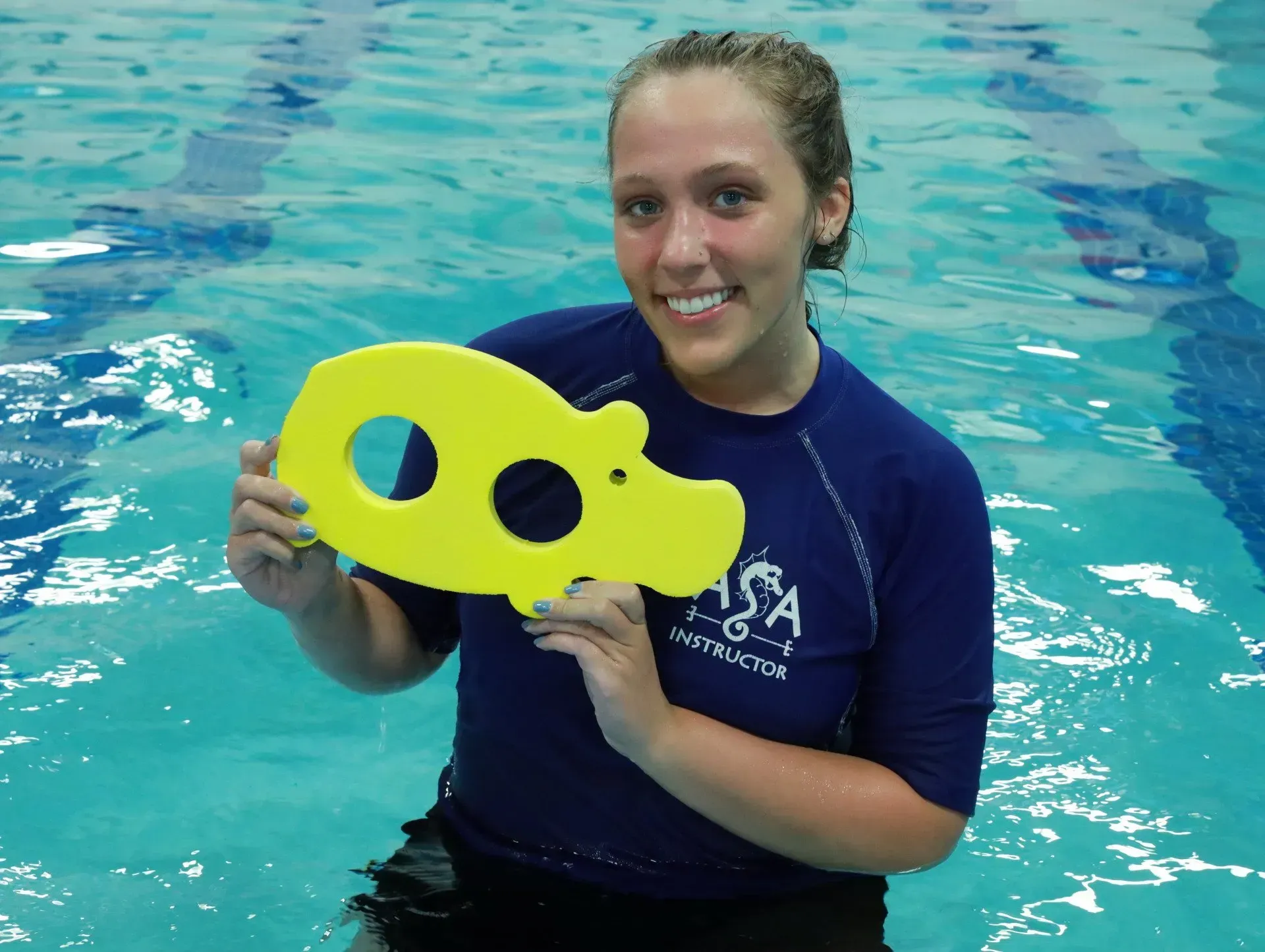 Woman in pool holding a yellow hippo-shaped swimming aid, smiling.