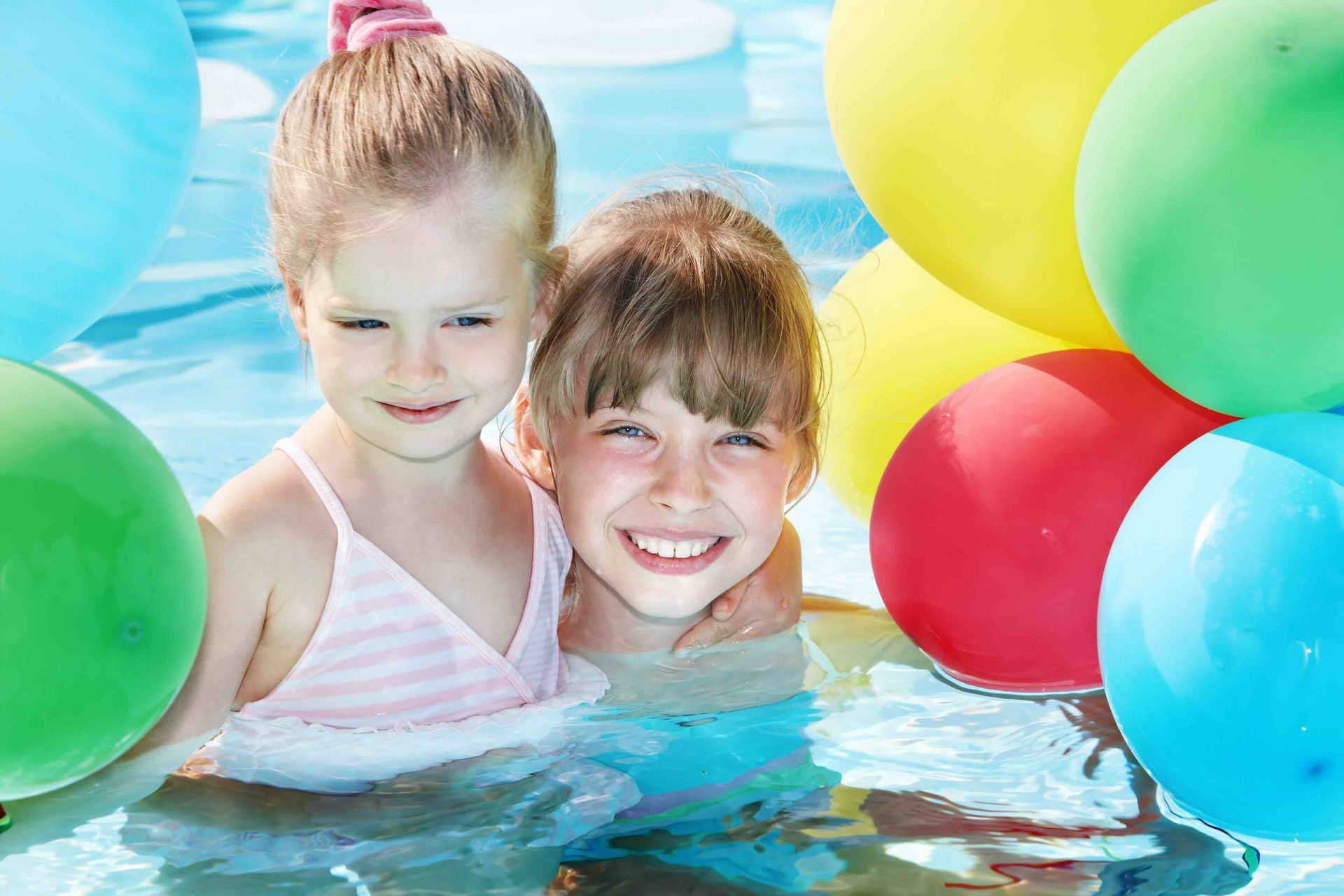 Two girls smiling in a pool, surrounded by colorful balloons.