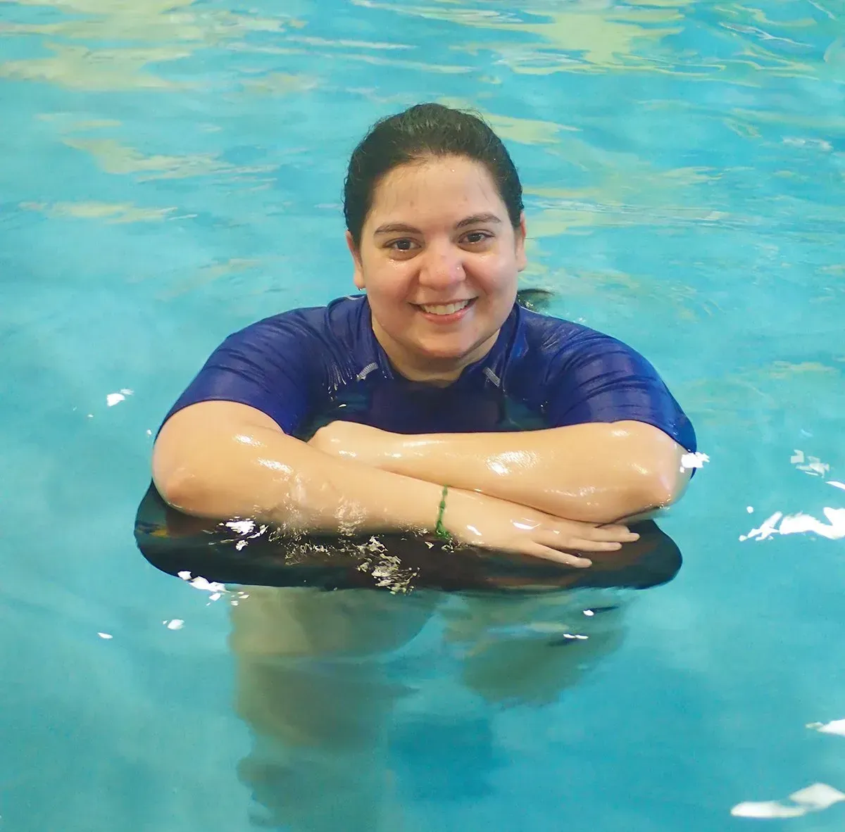 Woman smiling in pool, resting arms on black flotation device, wearing blue shirt.