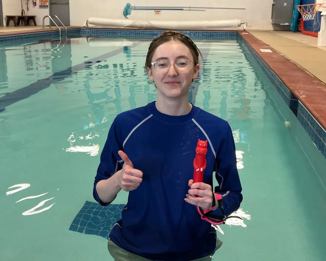 Person in a pool gives a thumbs up, holding a red whistle. Wearing a blue shirt and glasses.