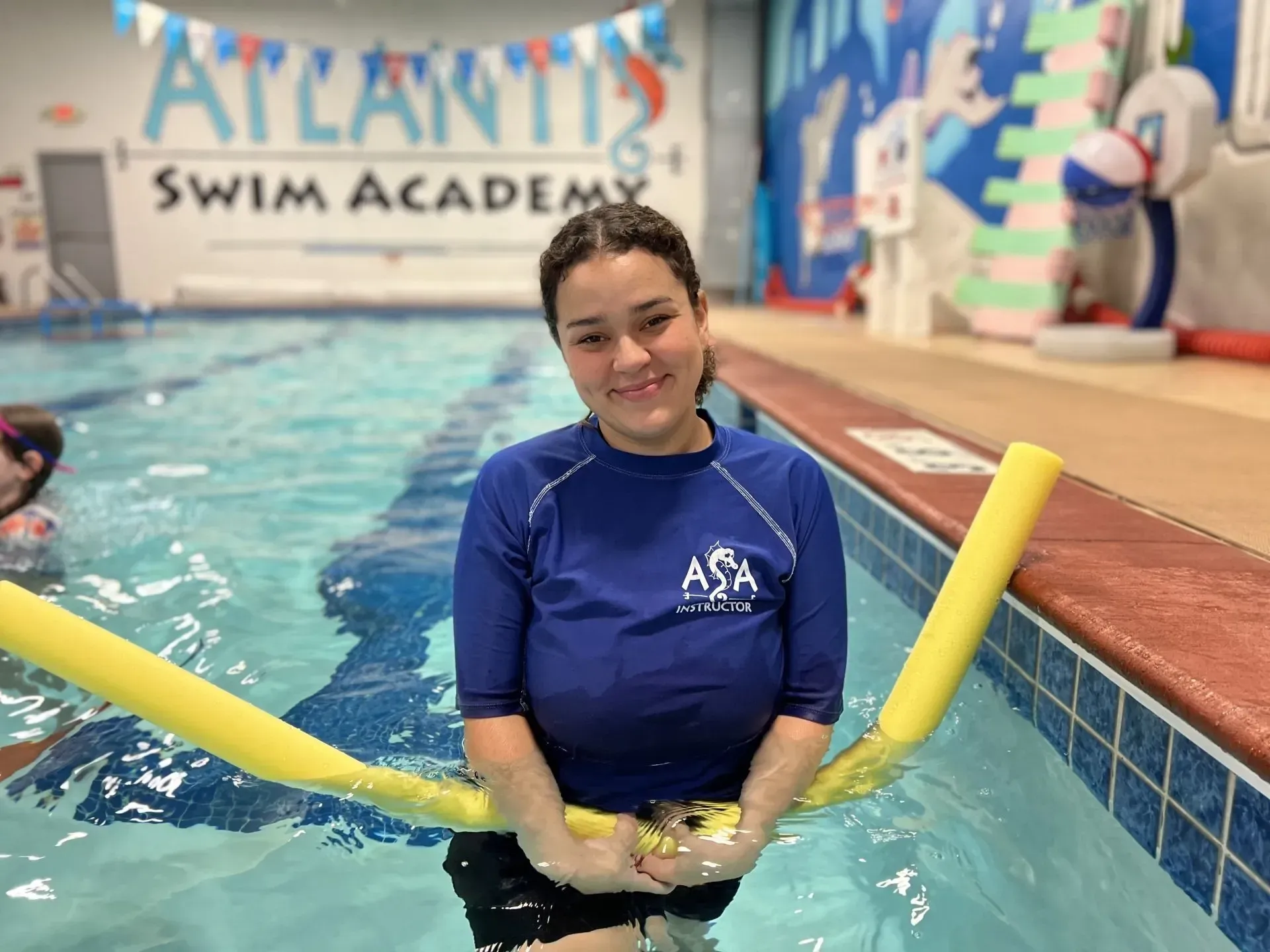 Woman in a blue shirt with a pool noodle in a pool at Atlantis Swim Academy smiles.