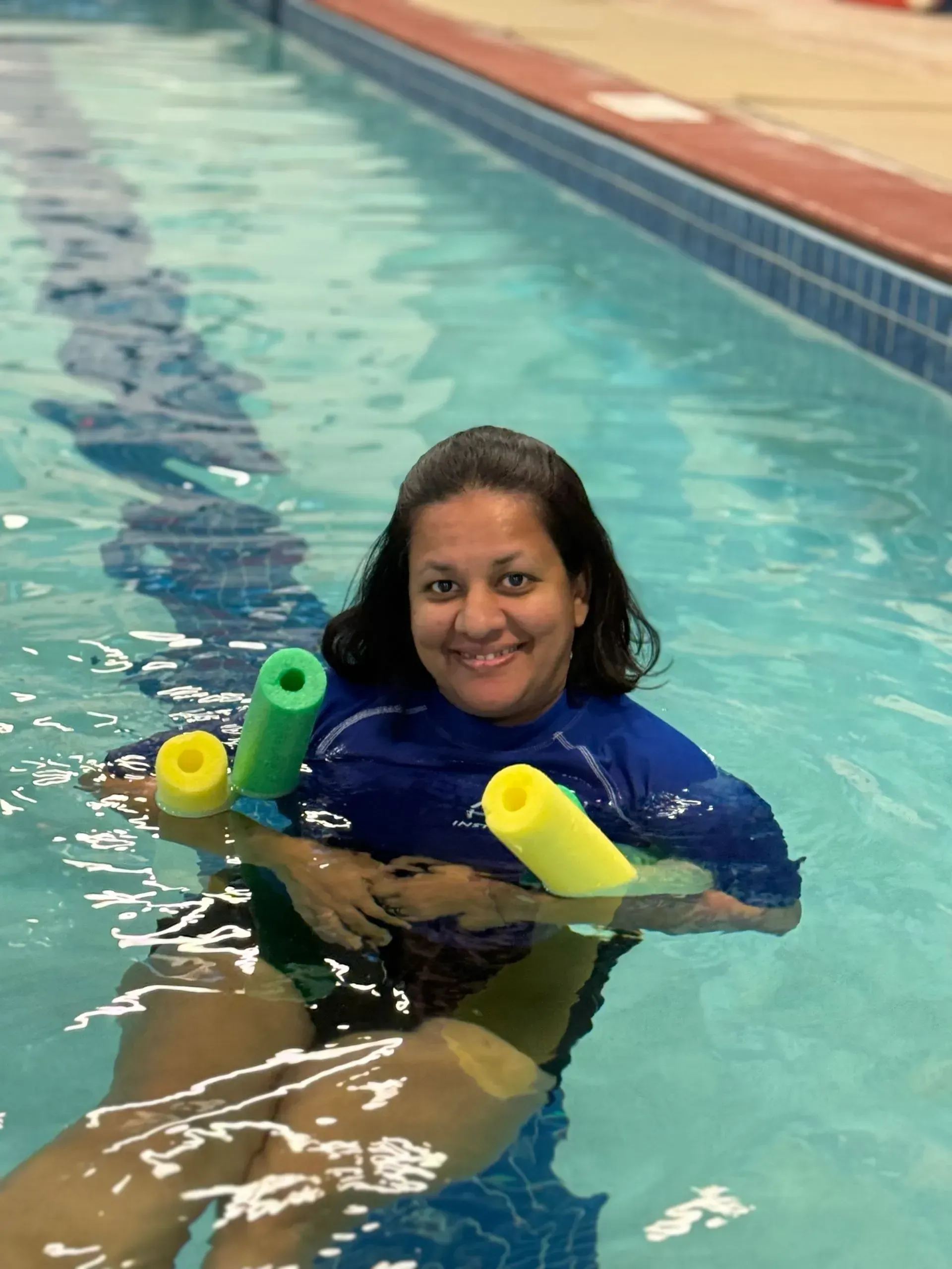 Woman in a pool wearing blue shirt, smiles while holding two pool noodles.
