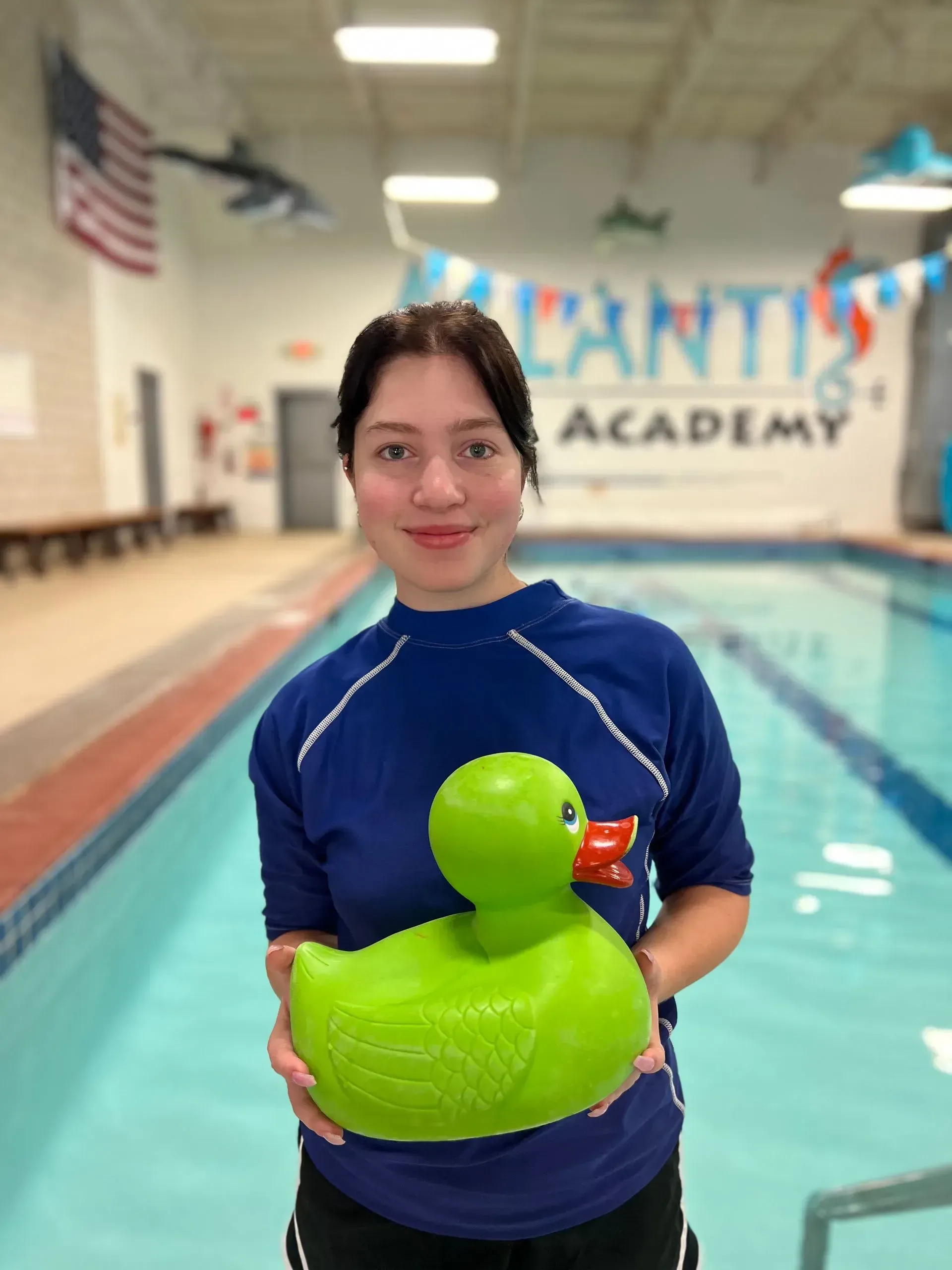Woman in blue shirt holds a large green duck near a pool; the backdrop is an indoor swimming pool.