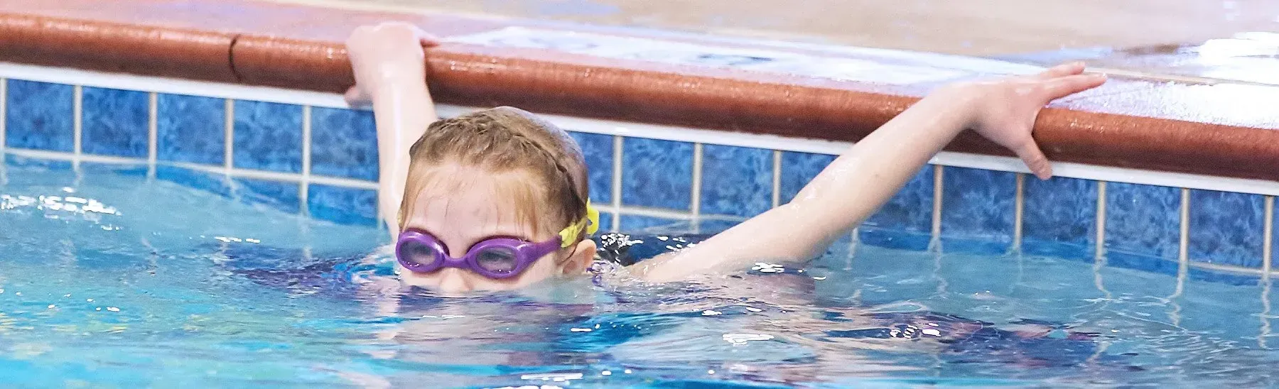 A person in purple goggles in a swimming pool, holding onto the edge.