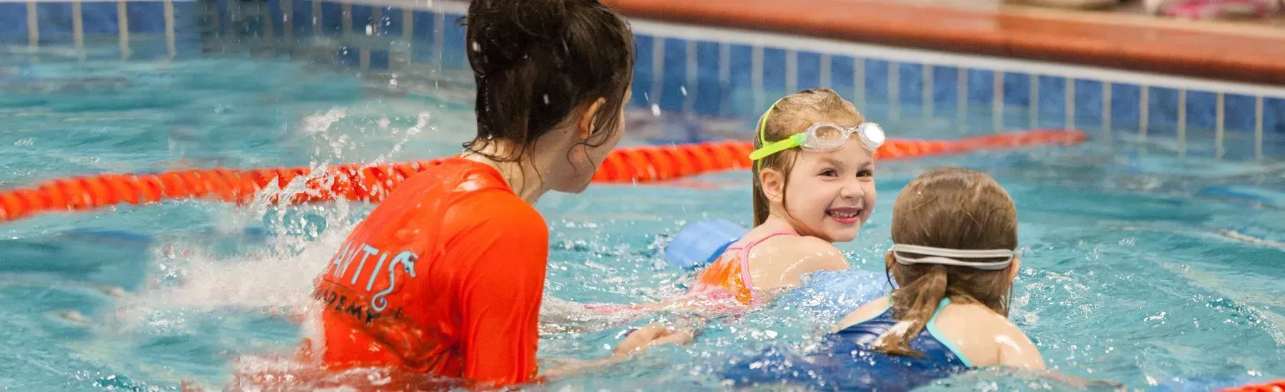 Three children learning to swim in a pool. One child smiles, wearing goggles. An instructor wears an orange shirt.
