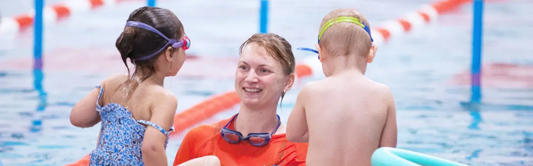 A woman smiles at two children in a pool. One child wears goggles, the other has their back to the camera.