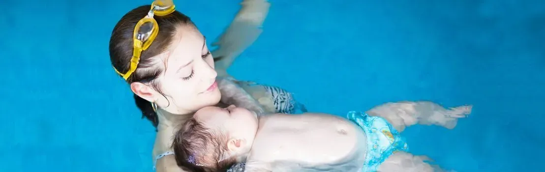 Woman with goggles holds a baby in a swimming pool. They are underwater.