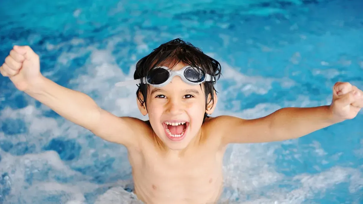 Boy in a pool with goggles, smiling and raising arms in excitement.
