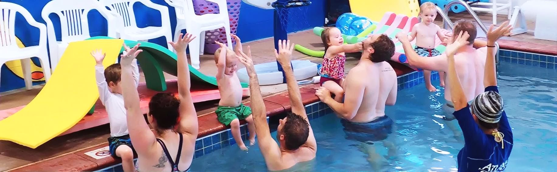 People in a swimming pool raise arms, with a yellow slide and a pool edge visible.