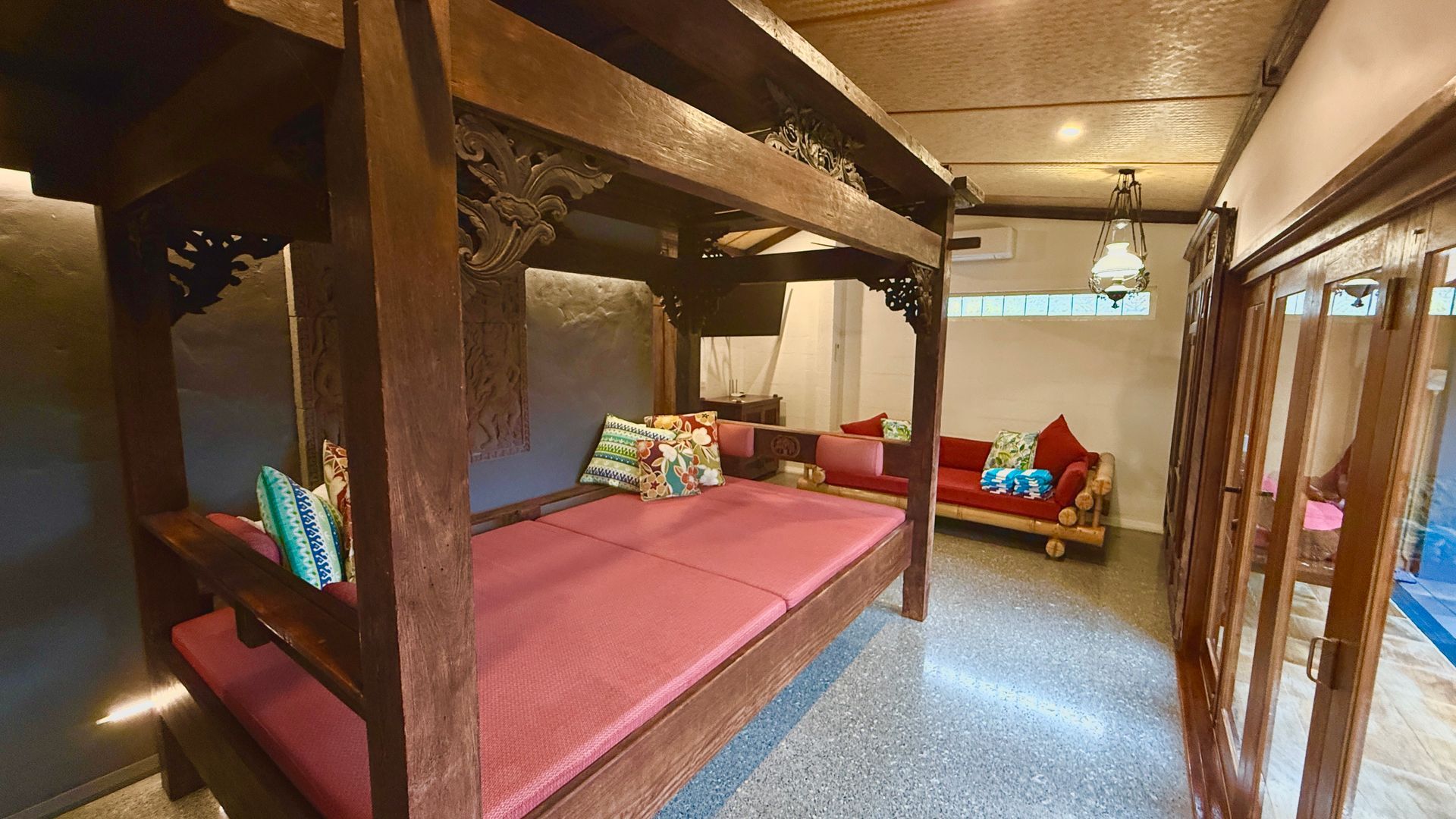 Bedroom with a large wooden bed, red mattress, and decorative pillows. A red sofa is in the background.