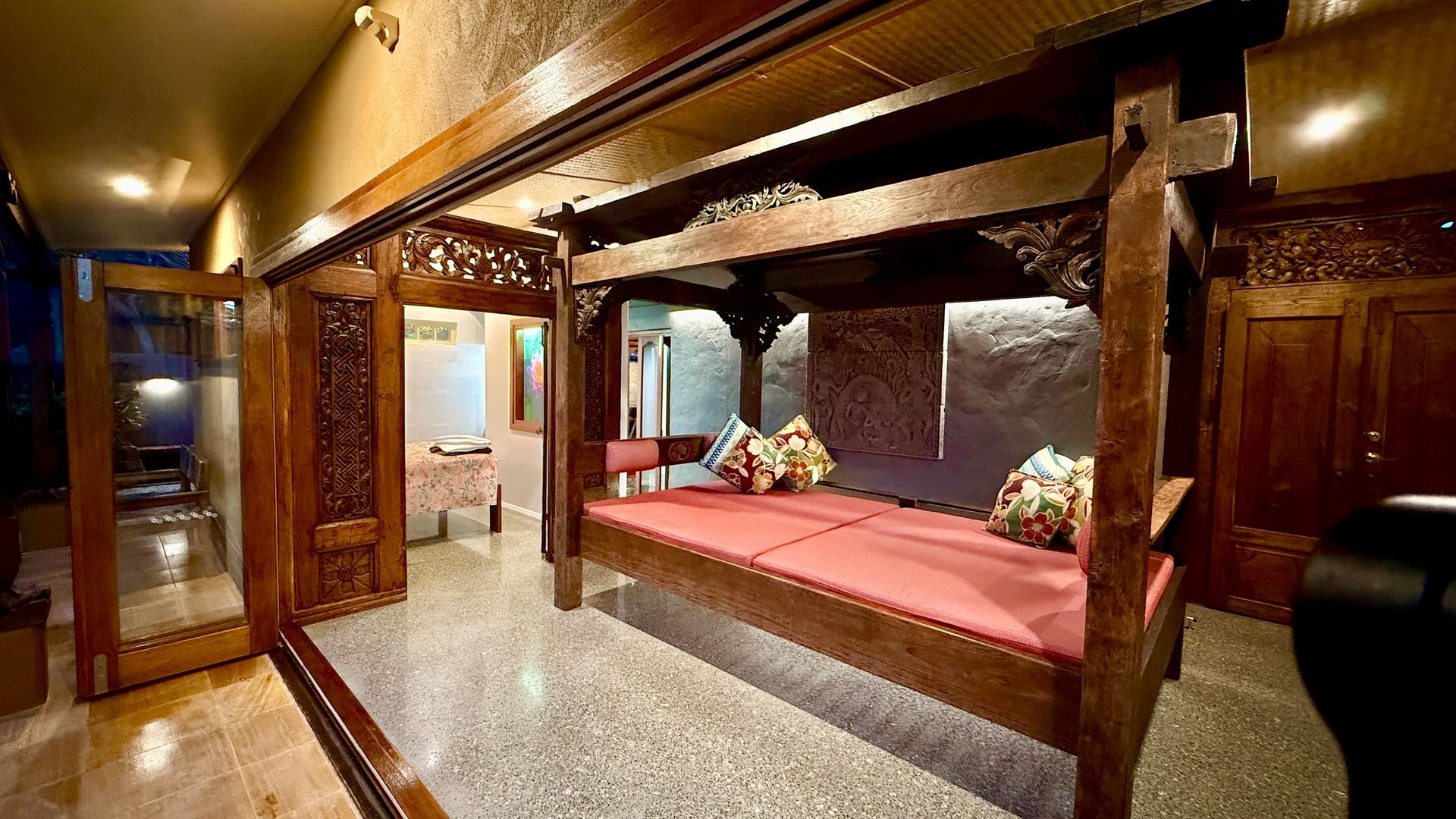 Ornate wooden four-poster bed in a warmly lit bedroom, with intricately carved doors and wall panels.