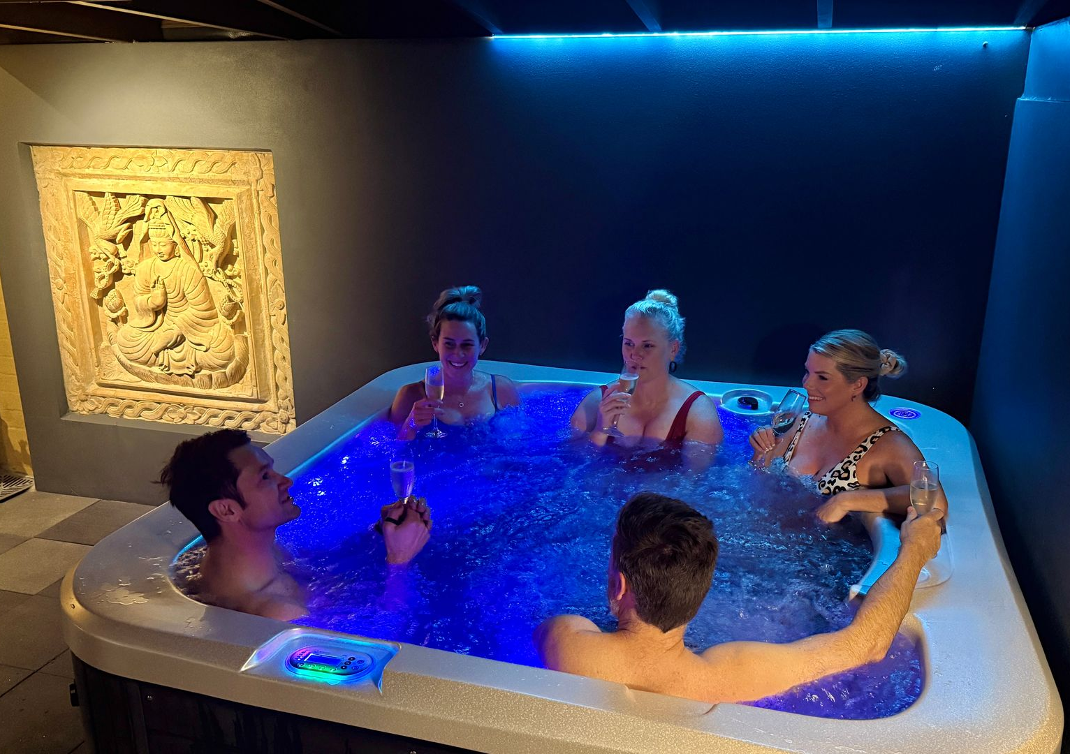 Group of friends in a hot tub, celebrating with drinks under blue lights.