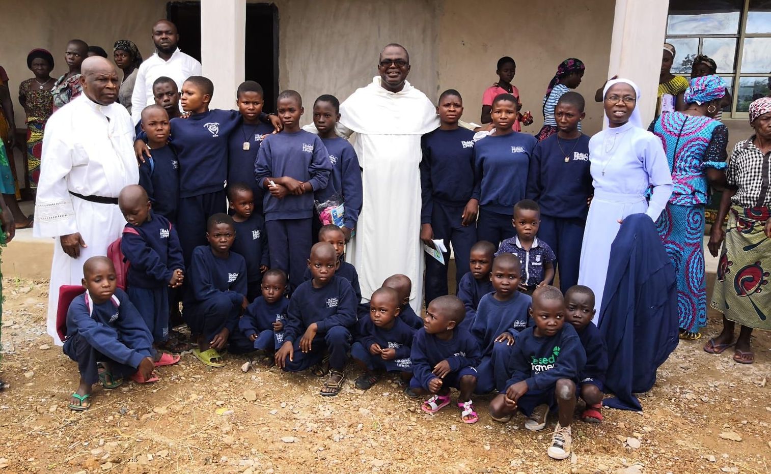 Nigerian school children standing around a Nigerian priest and nun and other clergy and local adults.