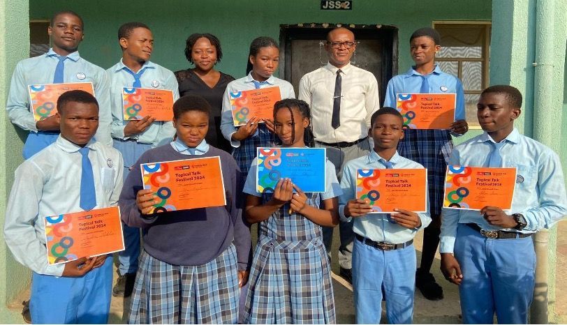 Group of students in school uniforms holding up certificates accompanied by their teachers