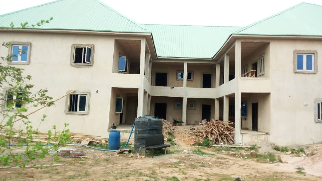 A two-story cream-colored building with a green roof under construction, surrounded by construction materials including wood planks and water containers in the foreground.