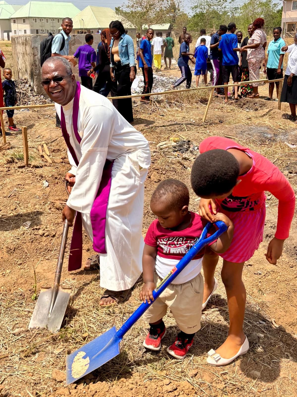 Smiling priest posing for camera and digging into soil, next to child helping small child/toddler hold a spade