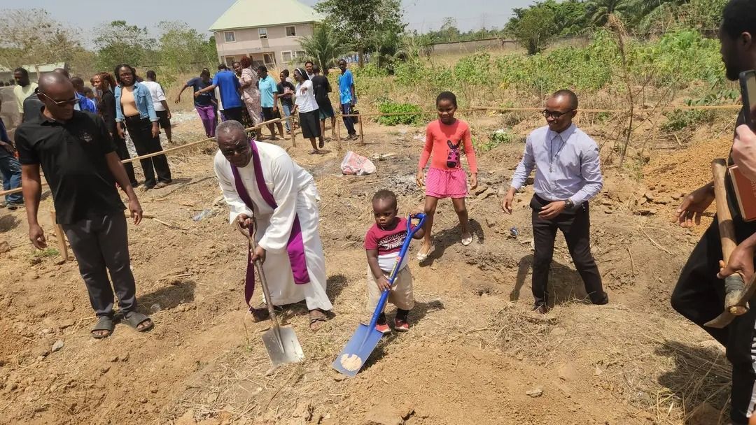 Priest and small African child holding shovels and digging into the ground, surrounded by community