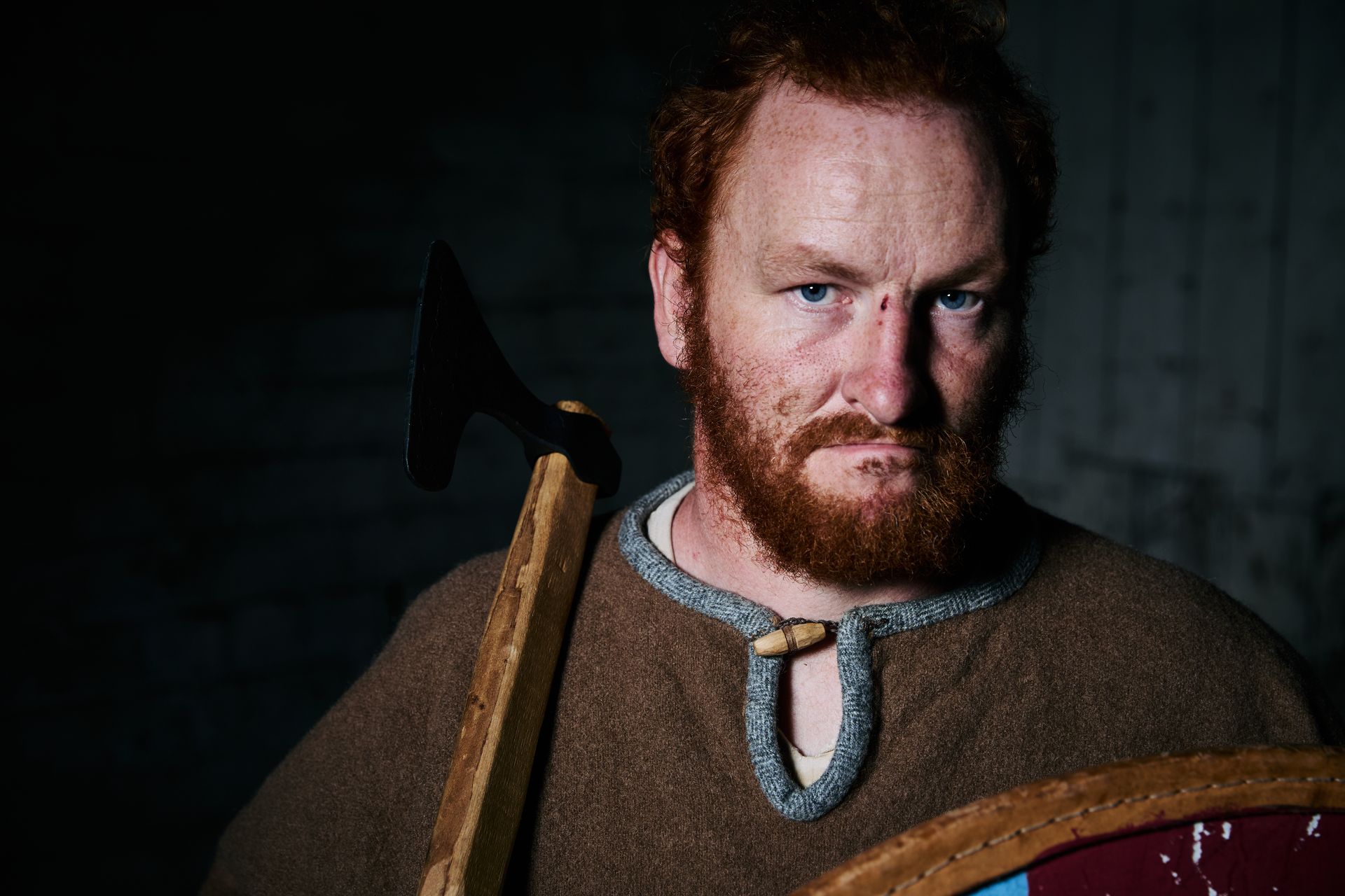 A bearded rugged man wearing viking clothes with an axe on his shoulder.