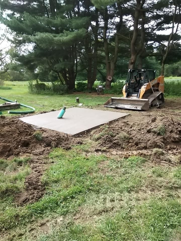 A small concrete pad in a yard being worked on by a skid steer. Earth is freshly dug, and grass surrounds the area.
