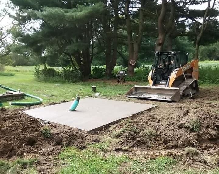 A concrete pad sits in a dirt clearing, with a skid-steer loader nearby.