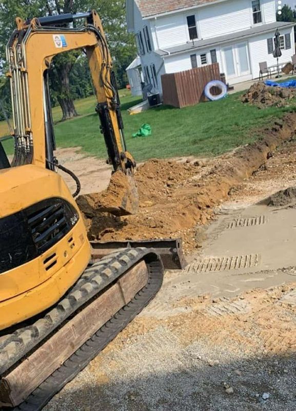 Yellow excavator digging earth in a yard. In the background, a house and other construction materials are visible.