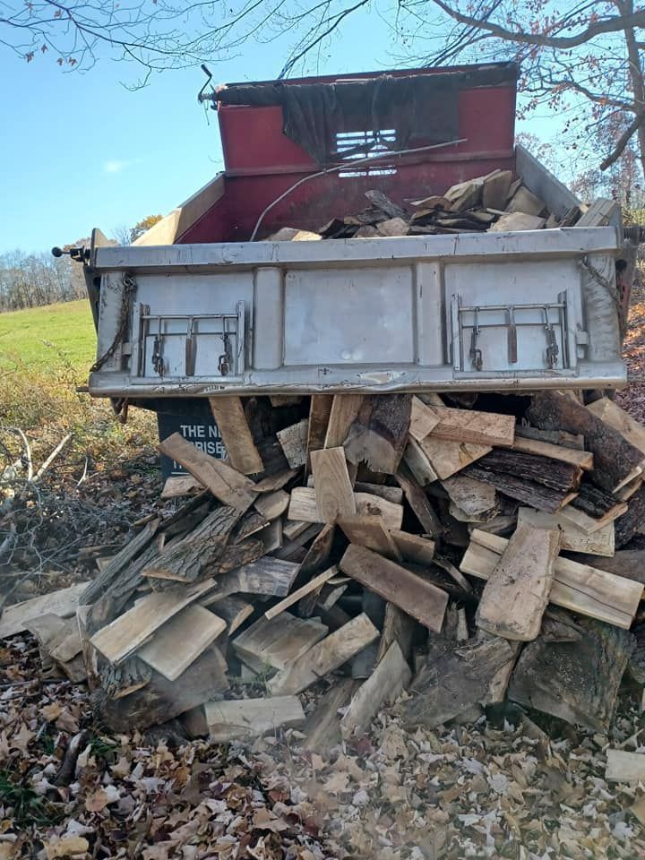 Truck bed overflowing with firewood; a red cab is visible in the background, grassy field beyond.