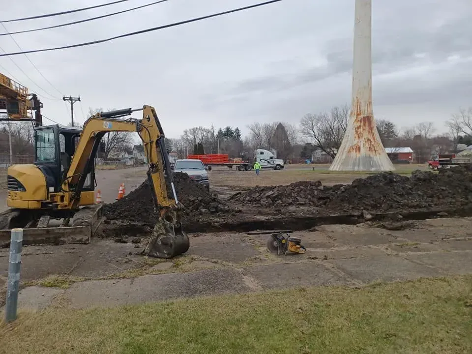 A yellow excavator digs a trench near a water tower and trucks on an overcast day.