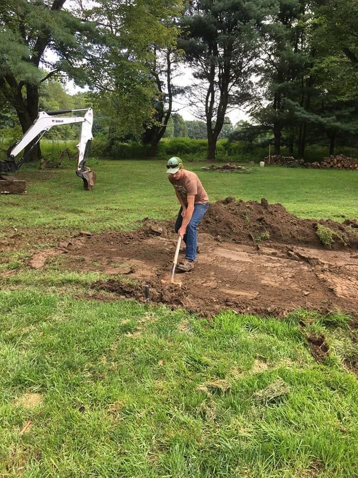 Man digging with a shovel in a yard with a Bobcat excavator in the background. Green grass and dirt mounds are visible.