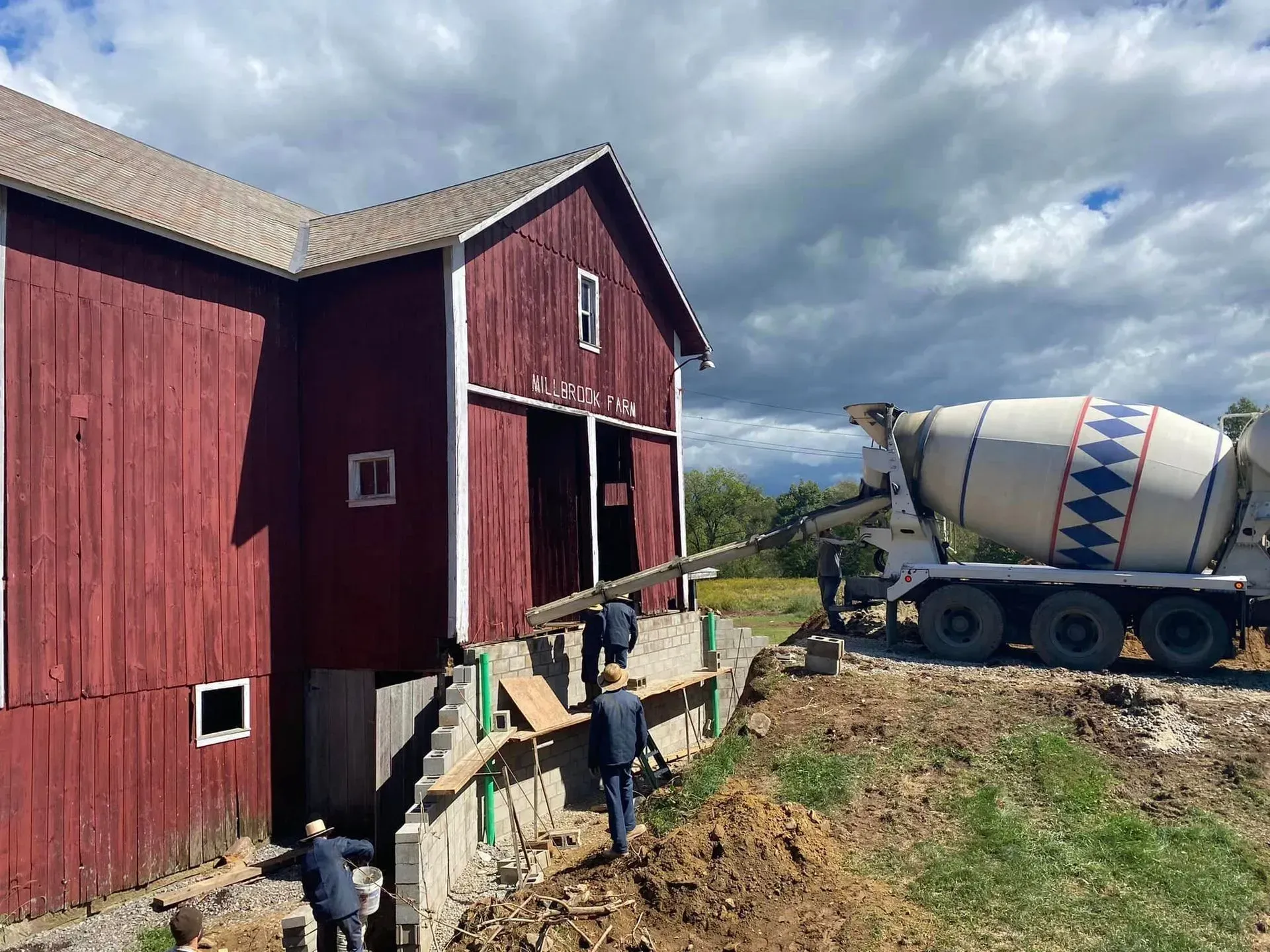 Cement truck pouring concrete into a new foundation under a red barn. Workers in dark clothing are assisting.