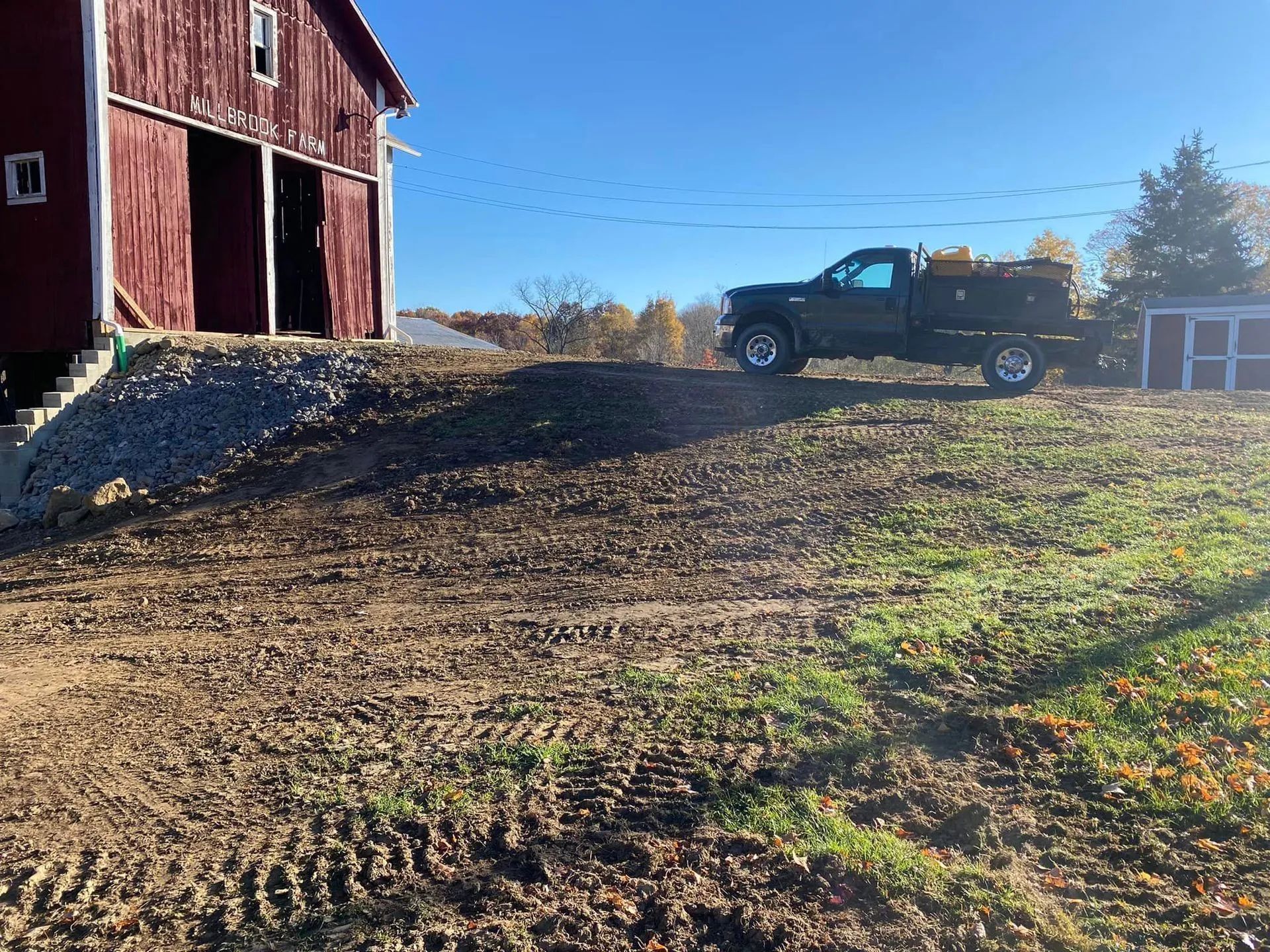 A red barn with open doors, a truck parked on a hill. Dirt and gravel are in the foreground.