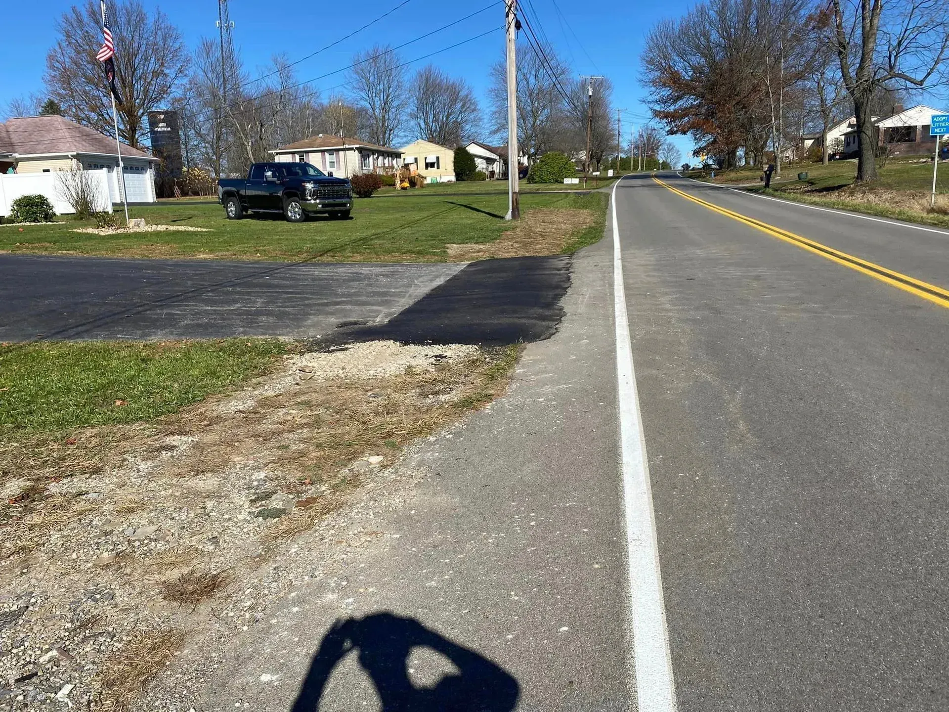 A street with a white line on the right side, a grassy area, and houses. A black pickup truck is parked.
