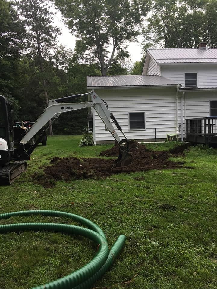 A Bobcat excavator digs in a backyard near a white house with a metal roof. Green corrugated pipe lies on the grass.