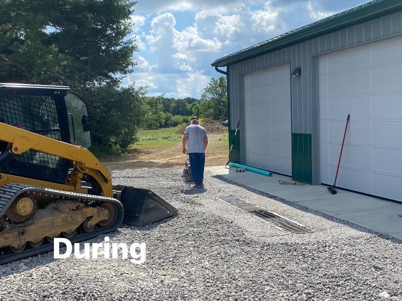A man using a compactor on a gravel driveway near a gray garage with green trim. A yellow skid steer is on the left.