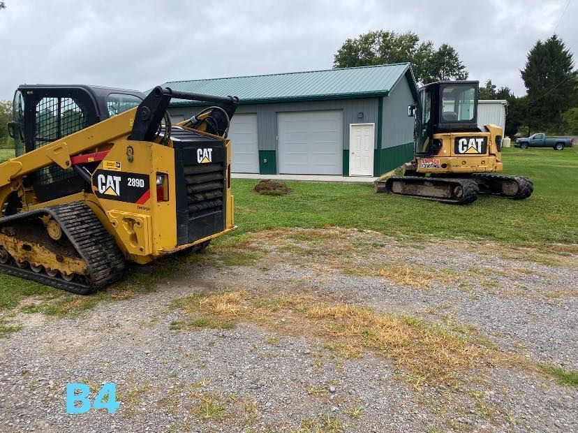 Two yellow CAT construction vehicles in front of a green-roofed building with two garage doors, in a grassy yard.