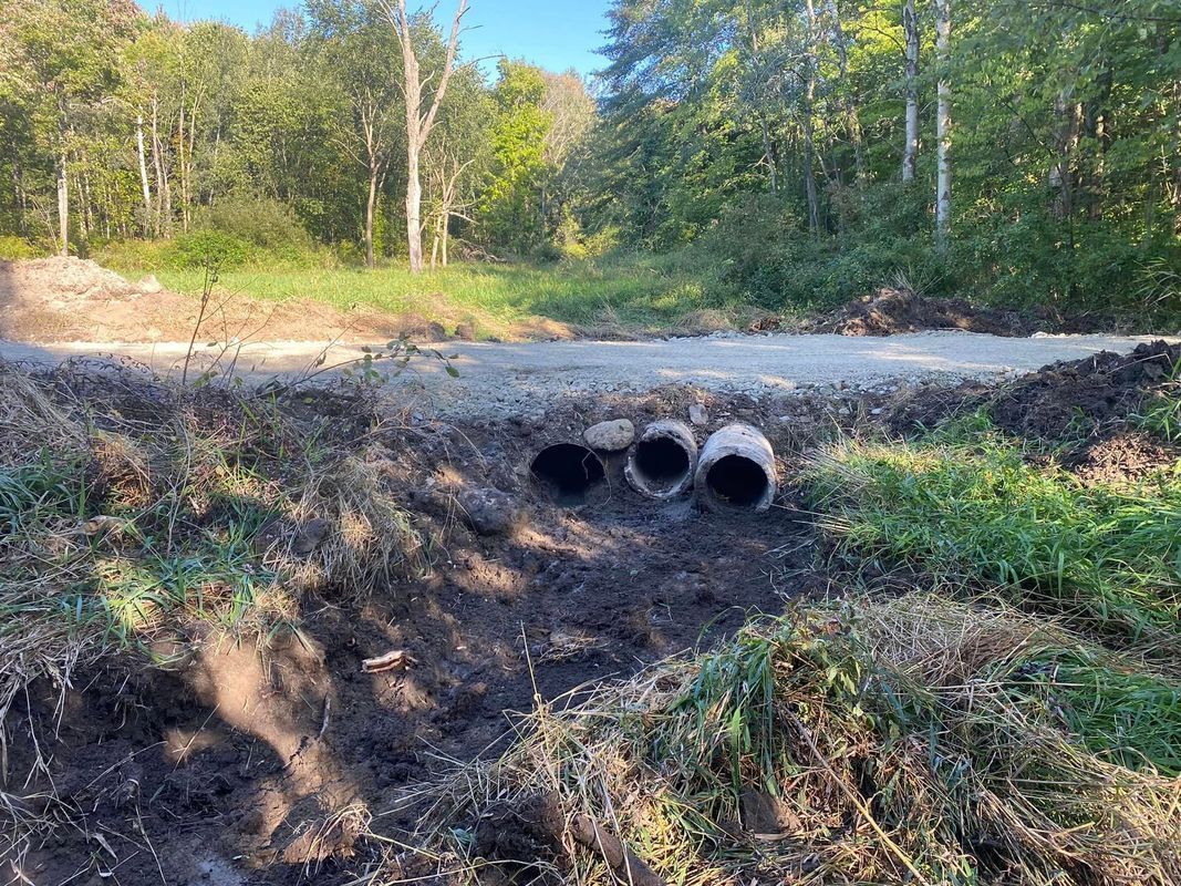 Three concrete pipes, partially buried, lie in a ditch under a gravel road. Trees and foliage are in the background.