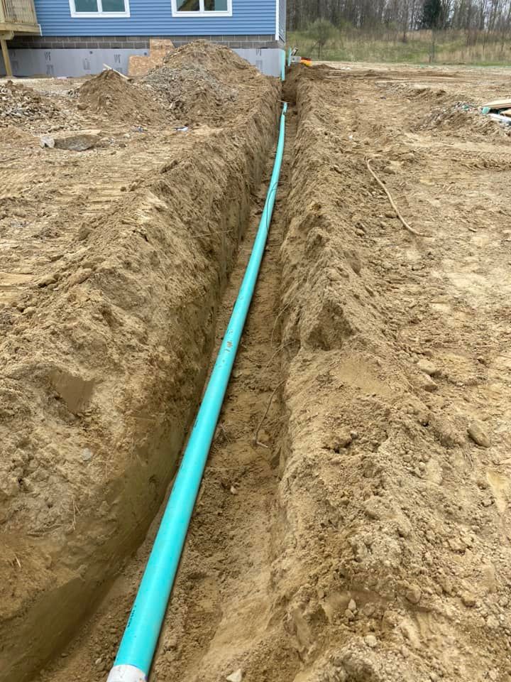 A trench dug in dirt holds a long, turquoise pipe. Construction site next to a blue house.
