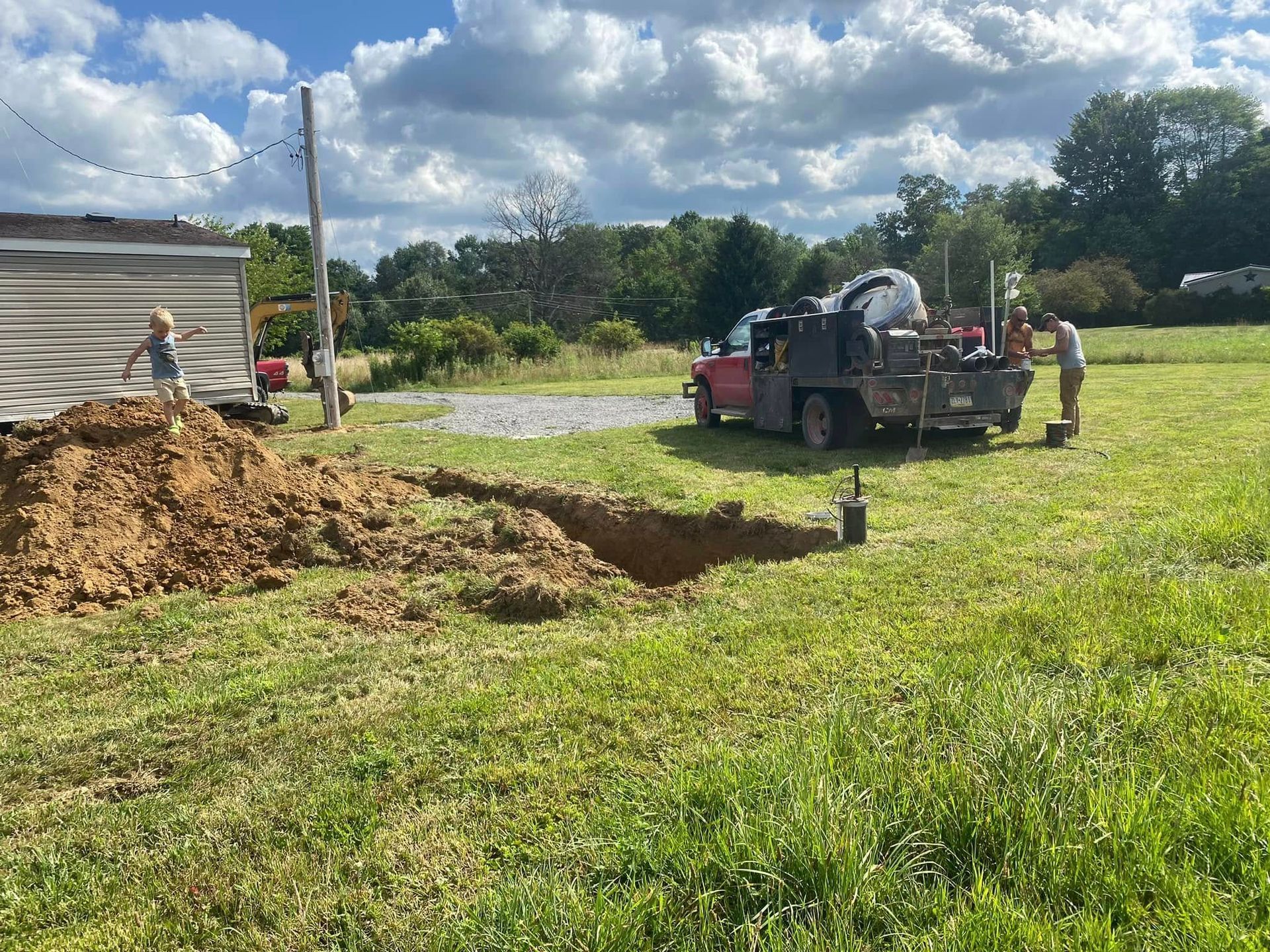 A red truck with equipment and two people working in a grassy field with a dug pit and a small child.
