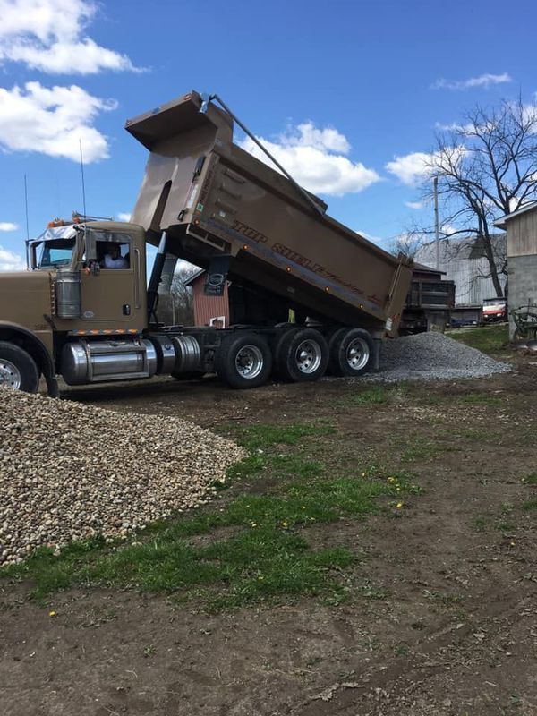 A brown dump truck is unloading gravel onto a pile on a grassy area, with a blue sky in the background.