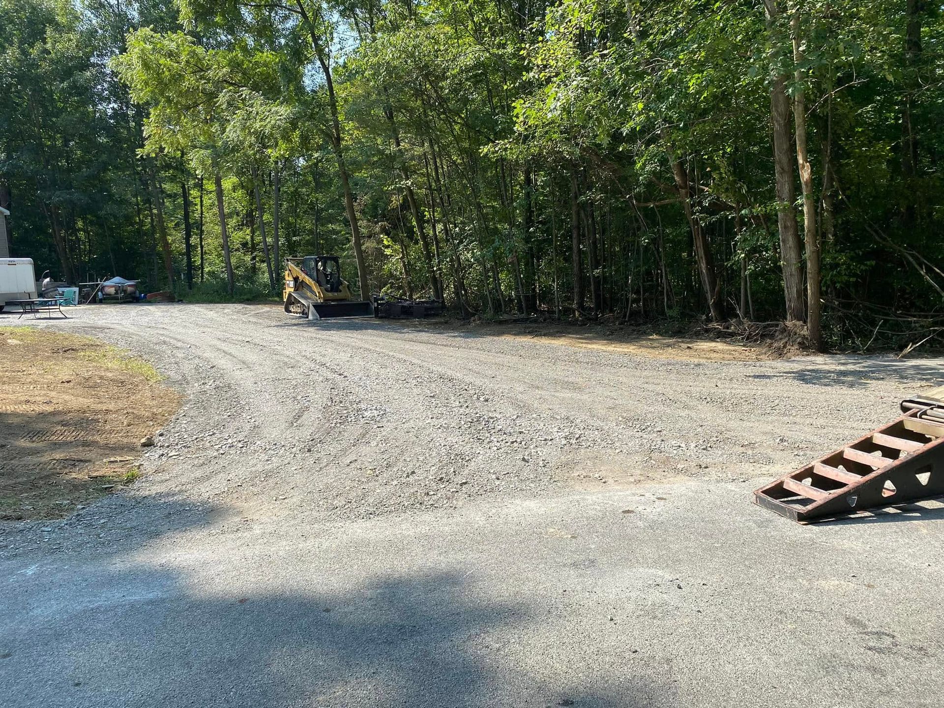Gravel driveway leading towards a wooded area with construction equipment and a ramp on the right.