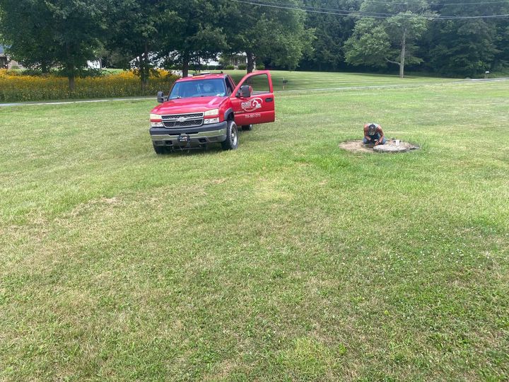Red pickup truck with open door parked on grassy field near a small, stone-ringed structure.