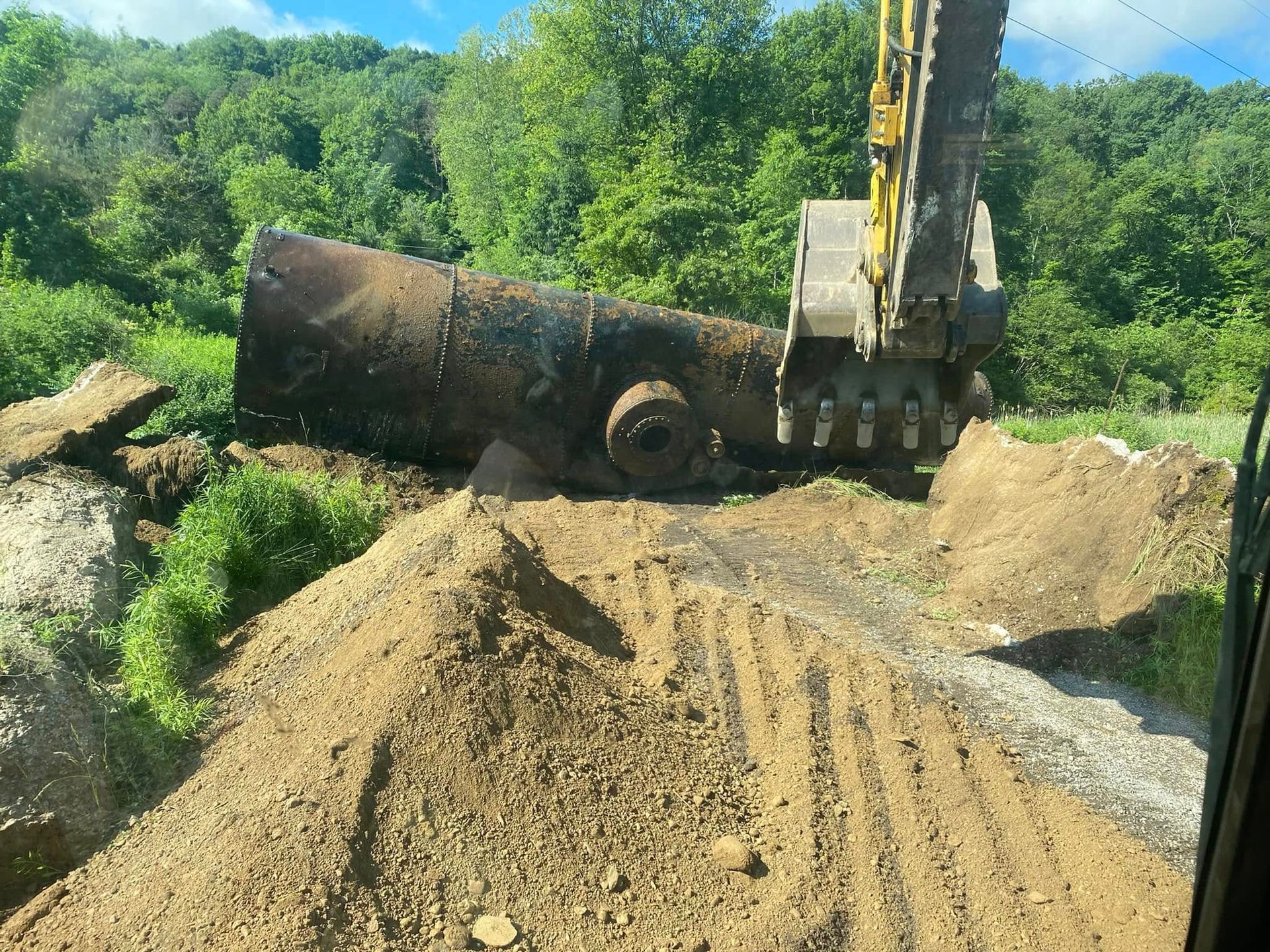 An excavator lifts a large, rusted metal tank from a pile of dirt in a green, wooded setting.